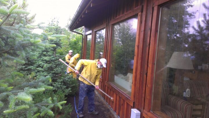 Painters in yellow raincoats and hats apply stain to the wooden exterior of a house surrounded by Portland's lush greenery.