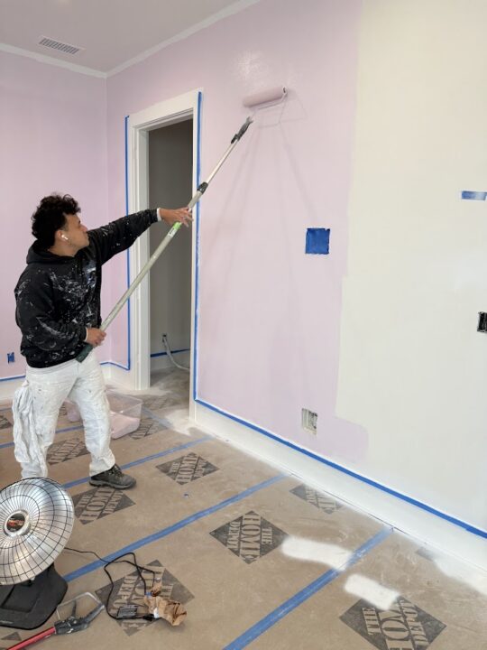 Painter rolls light pink paint on wall; protective sheets cover floor, blue painter's tape lines edges. Pearl Painters, Portland.